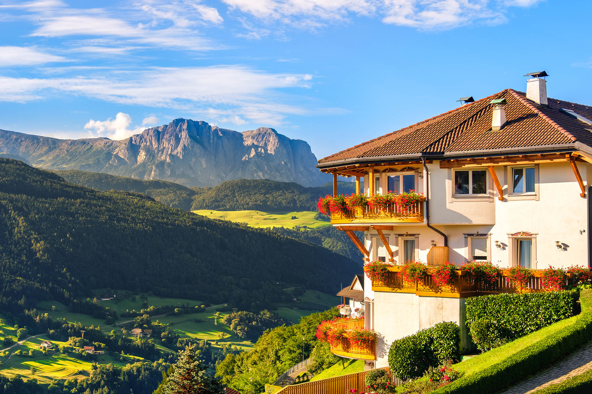Multistory building with balcony overlooks a sunny Italian Dolomites, with lush green surroundings.